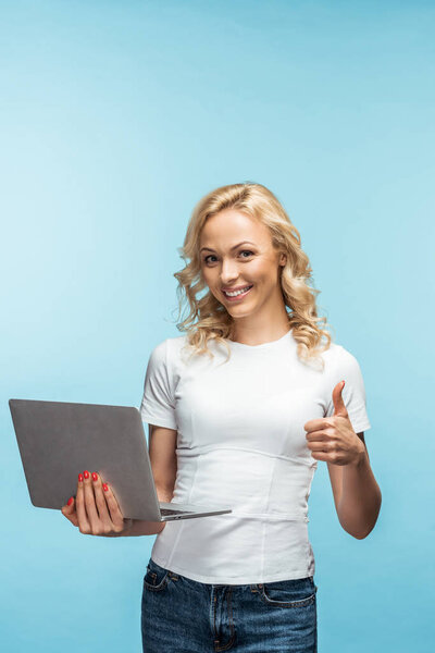 happy curly blonde woman holding laptop while showing thumb up on blue 