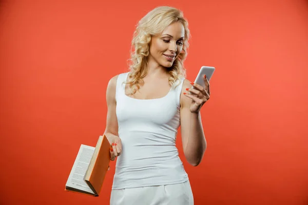 Alegre Mujer Rubia Sosteniendo Libro Mientras Utiliza Teléfono Inteligente Rojo — Foto de Stock