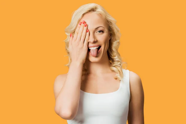 Šťastná Blondýnka Pokrývající Obličej Ukazující Jazyk Izolovaný Oranžový — Stock fotografie