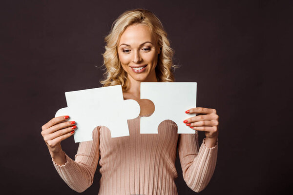 happy blonde woman connecting jigsaw pieces isolated on black 