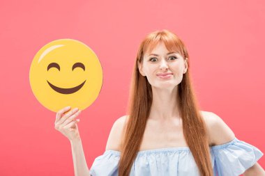 KYIV, UKRAINE - MAY 23, 2019: front view of cheerful redhead girl holding smiley isolated on pink clipart
