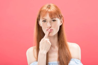 front view of curious redhead girl looking at camera and touching lips with finger isolated on pink clipart