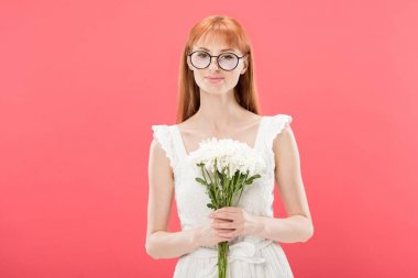 front view of attractive redhead girl in glasses and white dress holding flowers and smiling isolated on pink clipart
