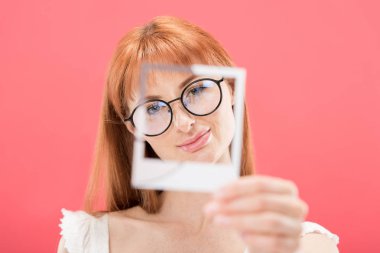 KYIV, UKRAINE - MAY 23, 2019: selective focus of attractive redhead girl in glasses holding vintage camera frame isolated on pink clipart