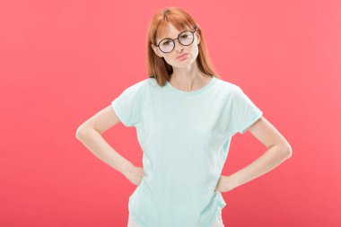 front view of curious redhead woman in glasses with hands on hips looking at camera isolated on pink clipart