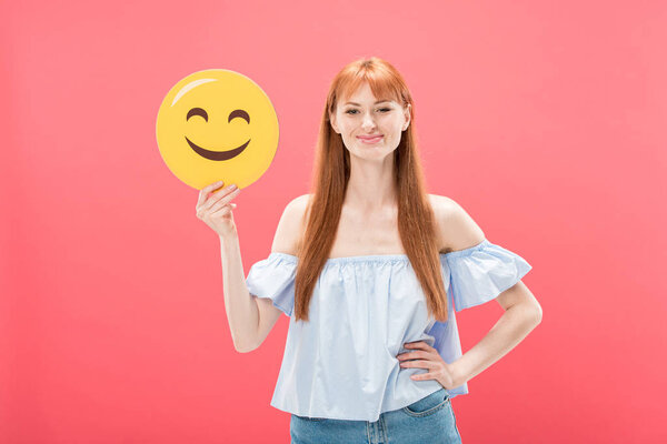 KYIV, UKRAINE - MAY 23, 2019: front view of redhead girl standing with hand on hip and holding smiley isolated on pink