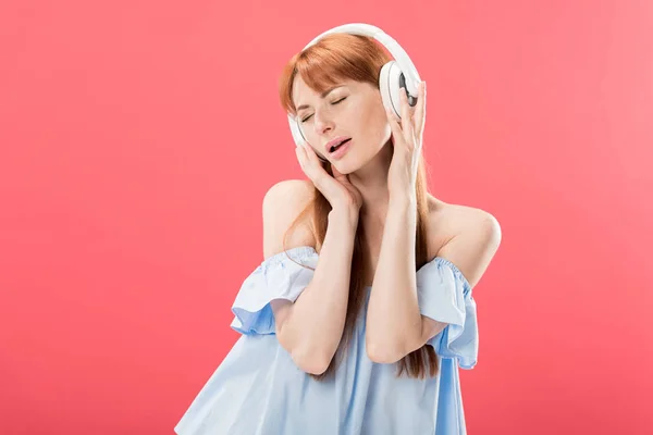 ピンクに隔離された目を閉じたヘッドフォンで音楽を聴く魅力的な赤毛の女性 — ストック写真