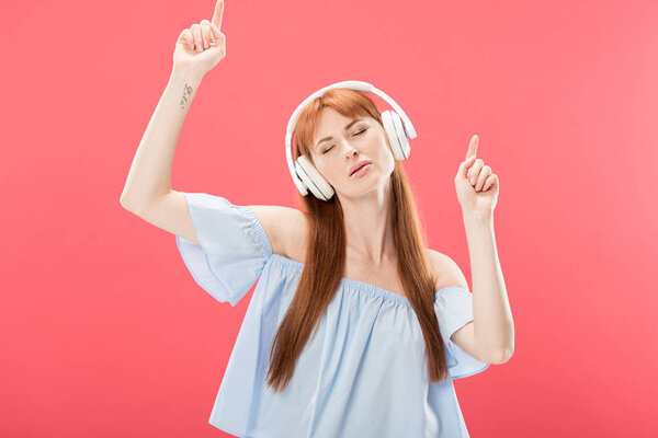 attractive redhead woman listening music in headphones with closed eyes and dancing isolated on pink