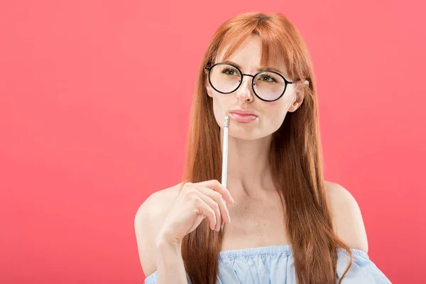 沉思红发年轻女子在眼镜拿着铅笔隔离在粉红色 — 图库照片