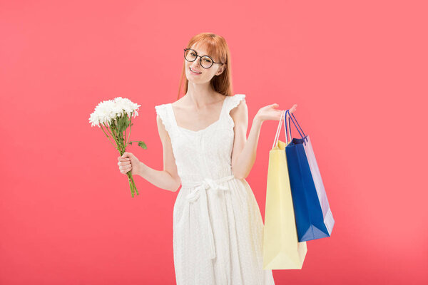 smiling redhead girl in glasses and white dress holding bouquet of flowers and shopping bags isolated on pink
