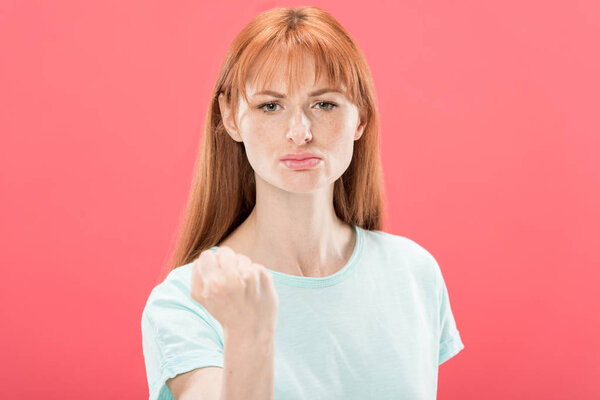 front view of displeased redhead woman in t-shirt showing fist and looking at camera isolated on pink