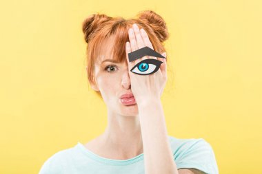 front view of redhead girl holding paper eye and eyebrow and looking at camera isolated on yellow clipart