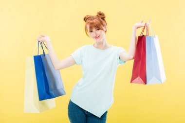 gülümseyen kızıl saçlı kız alışveriş çantaları tutan ve sarı izole kamera bakıyor
