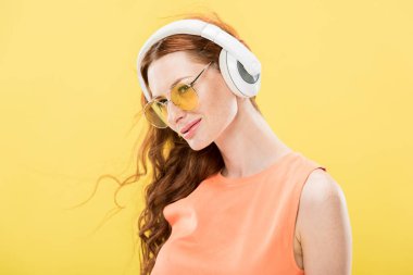güneş gözlüğü çekici kızıl saçlı kadın kulaklık müzik dinleme ve sarı izole gülümseyerek