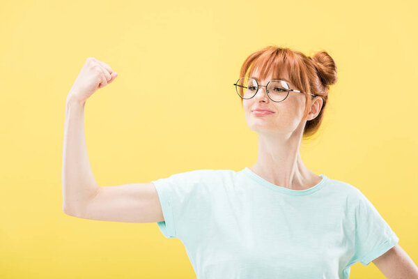 confident pleased redhead girl in glasses and t-shirt holding fist up isolated on yellow
