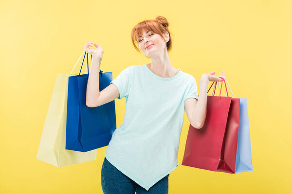 smiling redhead girl holding shopping bags with closed eyes isolated on yellow
