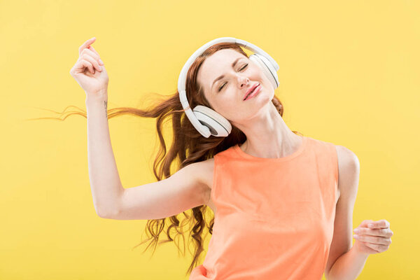 attractive smiling redhead woman listening music in headphones and dancing with closed eyes isolated on yellow