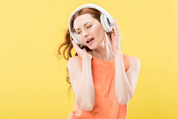 attractive redhead woman listening music in headphones and singing isolated on yellow