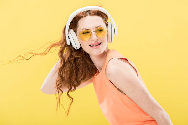 attractive redhead woman in sunglasses listening music in headphones and smiling isolated on yellow