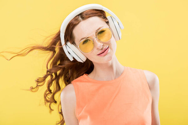 front view of attractive redhead woman in sunglasses listening music in headphones and smiling isolated on yellow
