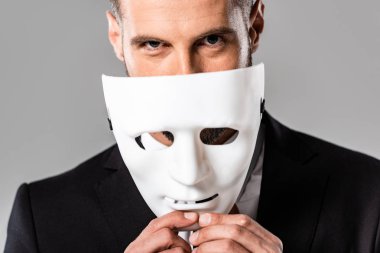 cunning businessman in black suit taking off white mask isolated on grey clipart