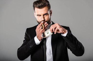 rich businessman in black suit lighting up cigar from burning dollar banknote isolated on grey clipart