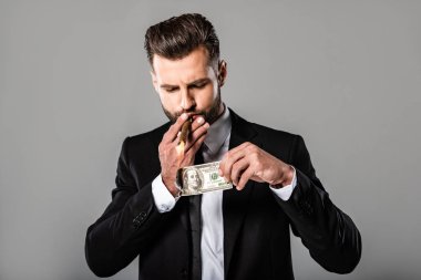 rich successful businessman in black suit lighting up cigar from burning dollar isolated on grey clipart