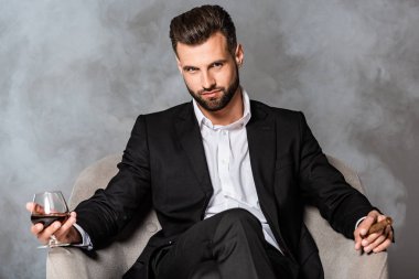siyah takım elbiseli kendinden emin işadamı duman puro ve viski ile koltukta oturan