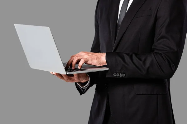 裁剪视图的商人在黑色西装使用笔记本电脑隔离在灰色 — 图库照片