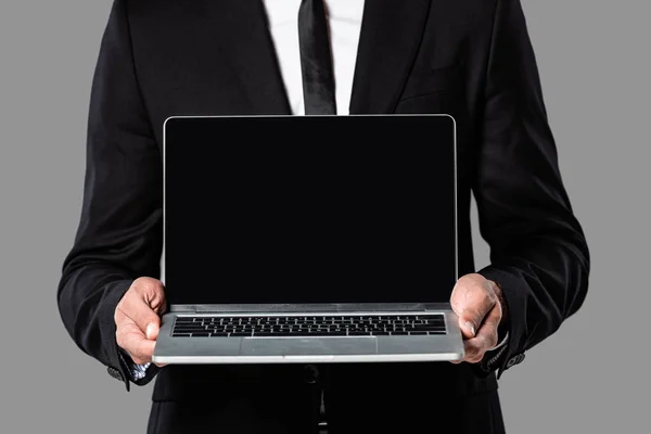 裁剪视图的商人在黑色西装显示笔记本电脑与空白屏幕隔离在灰色 — 图库照片