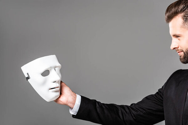 side view of smiling handsome businessman in black suit looking at white mask isolated on grey