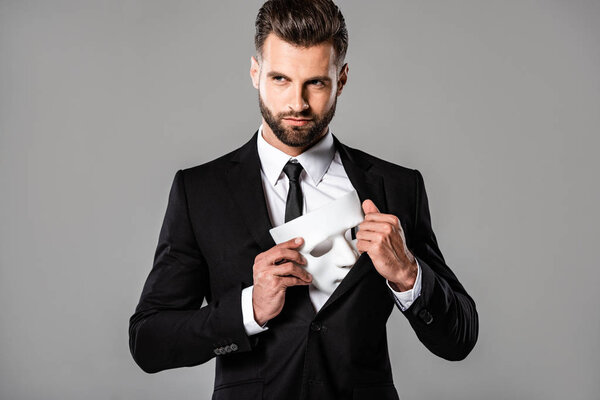 sly businessman in black suit hiding white mask isolated on grey