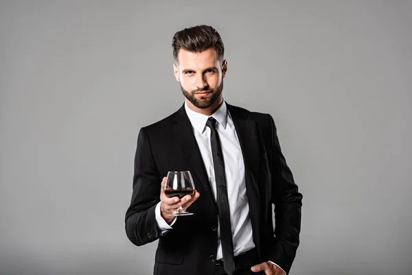 英俊的商人在黑色西装拿着玻璃与威士忌隔离在灰色 — 图库照片