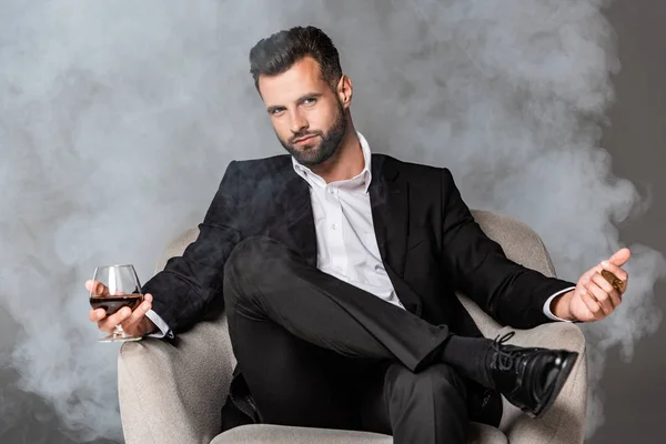 黒いスーツを着たビジネスマンが 煙の中で葉巻とウイスキーを持つアームチェアに座っている — ストック写真