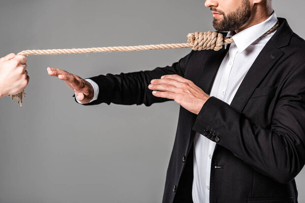 cropped view of businessman in black suit with noose on neck showing stop gesture to murderer with rope isolated on grey
