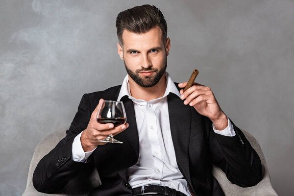 successful businessman in black suit sitting in armchair with cigar and whiskey in smoke