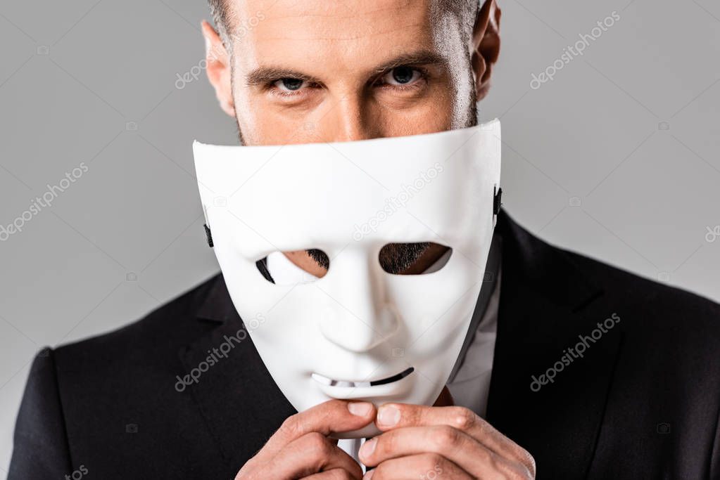 cunning businessman in black suit taking off white mask isolated on grey