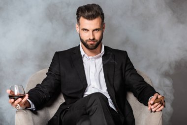 siyah takım elbiseli başarılı işadamı duman puro ve viski ile koltukta oturan