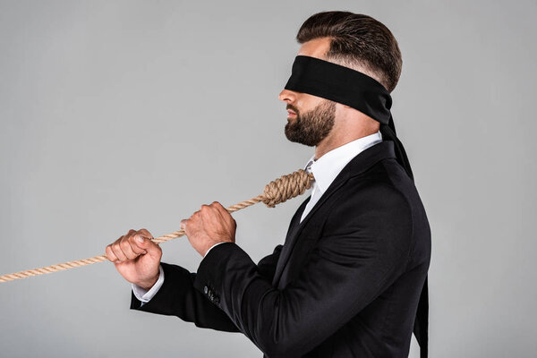 side view of blindfolded businessman in black suit with noose on neck holding rope isolated on grey