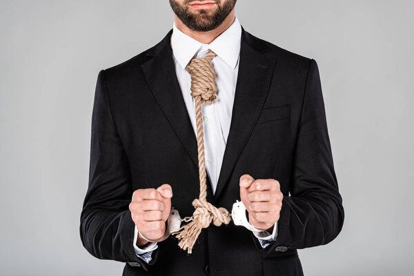 partial view of businessman in black suit and handcuffs with noose on neck isolated on grey