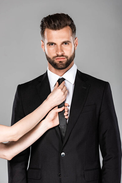 woman touching tie on confident businessman in black suit isolated on grey