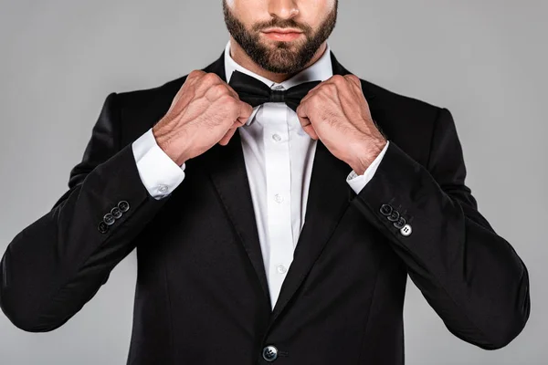 灰色で隔離された蝶ネクタイを固定する黒いスーツのエレガントな男のトリミングされたビュー — ストック写真