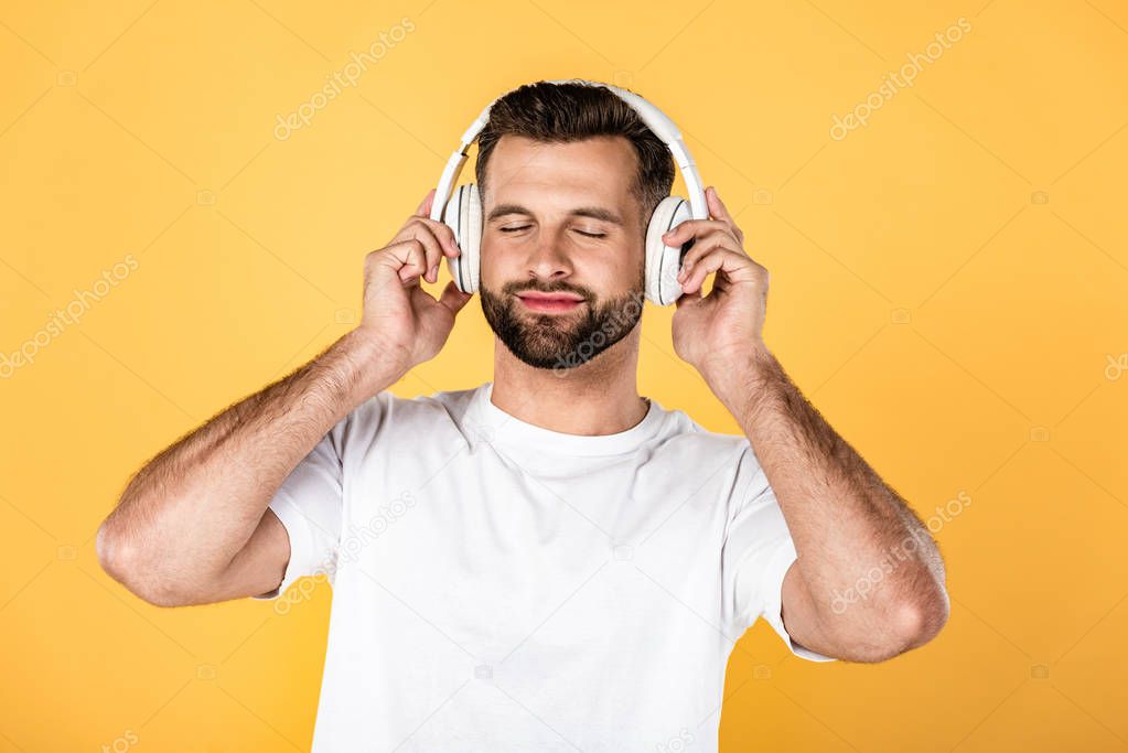 happy handsome man in white t-shirt listening music in headphones with closed eyes isolated on yellow