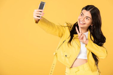 zafer işareti gösteren ve akıllı telefonda selfie alarak güzel kız, sarı izole