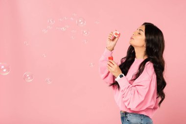 cheerful woman blowing soap bubbles isolated on pink clipart
