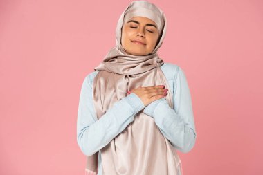 happy muslim girl holding hands on heart, isolated on pink clipart