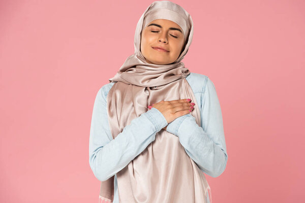 happy muslim girl holding hands on heart, isolated on pink