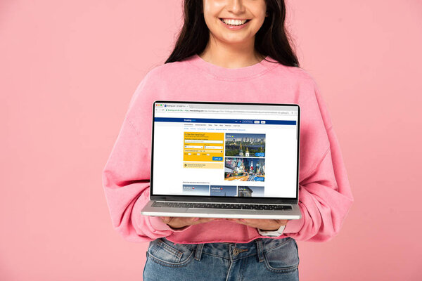 KYIV, UKRAINE - JULY 30, 2019: cropped view of smiling girl holding laptop with booking website on screen, isolated on pink
