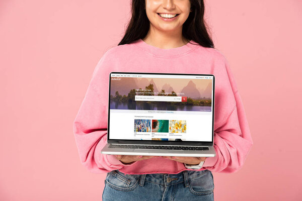 KYIV, UKRAINE - JULY 30, 2019: cropped view of smiling girl holding laptop with shutterstock website on screen, isolated on pink