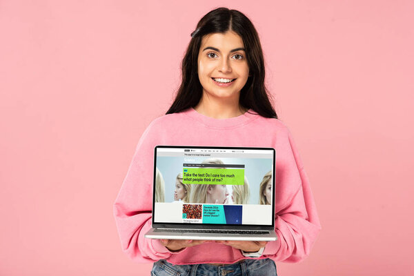 KYIV, UKRAINE - JULY 30, 2019: smiling girl holding laptop with BBC website on screen, isolated on pink